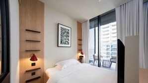 Quarto de hotel com uma cama, uma televisão montada na parede, um abajur vermelho, uma janela com vista para um edifício e uma porta.