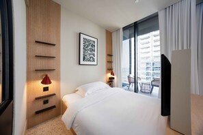 Ein Hotelzimmer mit einem Bett, einem an der Wand befestigten Fernseher, einer roten Lampe, einem Fenster mit Blick auf ein Gebäude und einer Tür.