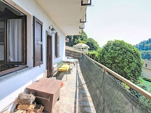 Un balcon avec une table et des chaises en bois, un mur blanc avec des fenêtres et une vue sur un paysage verdoyant.