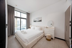 A modern hotel room with a large bed, a bedside table with a lamp, a dresser, and a view of the cityscape through large windows. - Modern 2BR Retreat | Near Downtown Dubai (Dubai)