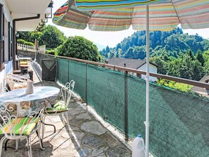 A balcony with a table and chairs, a striped umbrella, and a view of a forested area. - Apartment Susy by Interhome (Miazzina)