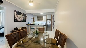 A dining area with a glass table surrounded by chairs, a ceiling fan, and a kitchen in the background. - Just 50 meters from the beach with 2 bedrooms and parking (Balneário Camboriú)