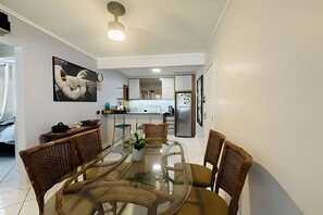 Un comedor con mesa de vidrio rodeada de sillas, un ventilador de techo y una cocina al fondo.