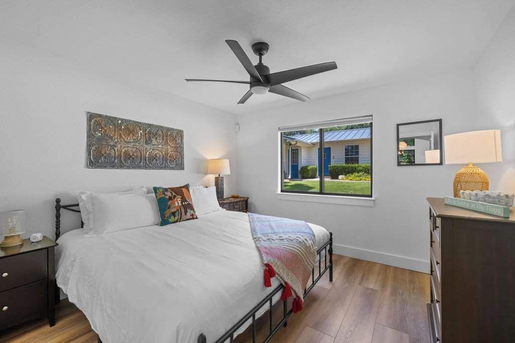 A bedroom with a bed, a ceiling fan, a window with a view, a bedside table, and a dresser.