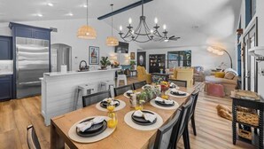 A modern kitchen with a central island, a dining area with a wooden table set for four, and a living space with a sofa and a dog. - Lakeshore Retreat | Pool, Spa & Private Peninsula (Kingsland)