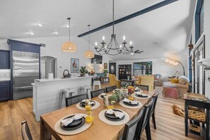 A modern kitchen with a central island, a dining area with a wooden table set for four, and a living space with a sofa and a dog. - Lakeshore Retreat | Pool, Spa & Private Peninsula (Kingsland)