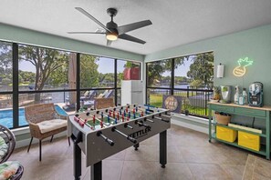 Ein Zimmer mit einem Kicker, einem Deckenventilator, großen Fenstern mit Blick auf einen Pool und Bäume, und einem Neon-Ananas-Schild.