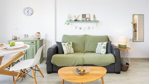 A living room with a green sofa, a wooden coffee table, a dining table with chairs, a clock on the wall, and a shelf with decorative items. - TenoHomes Atlantic View Icod Terrace (San Marcos)