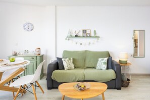 Ein Wohnzimmer mit einem grünen Sofa, einem Couchtisch aus Holz, einem Esstisch mit Stühlen, einer Wanduhr und einem Regal mit Dekoration.