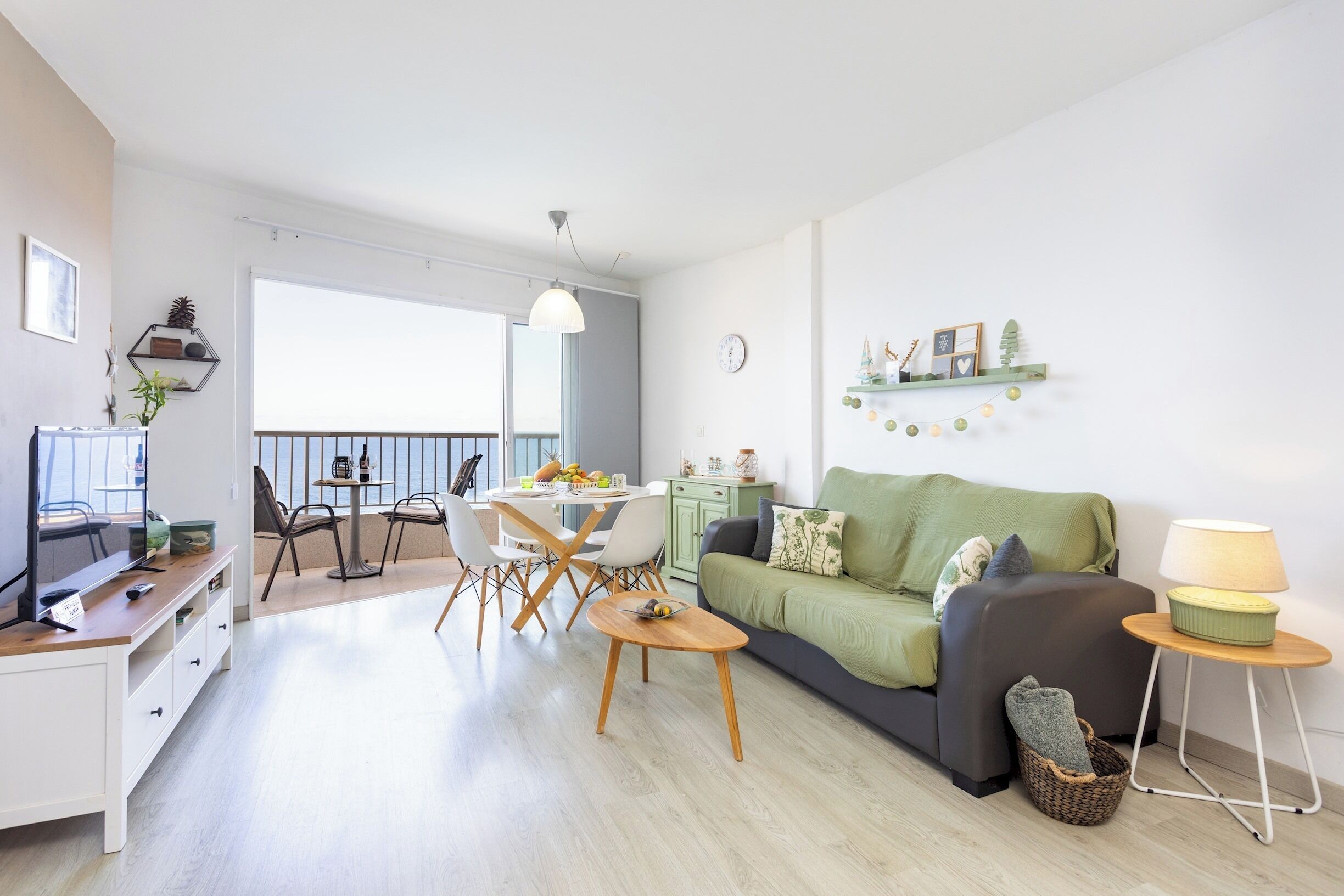 A modern living room with a green sofa, a dining area with a table and chairs, a TV, and a balcony with a view.