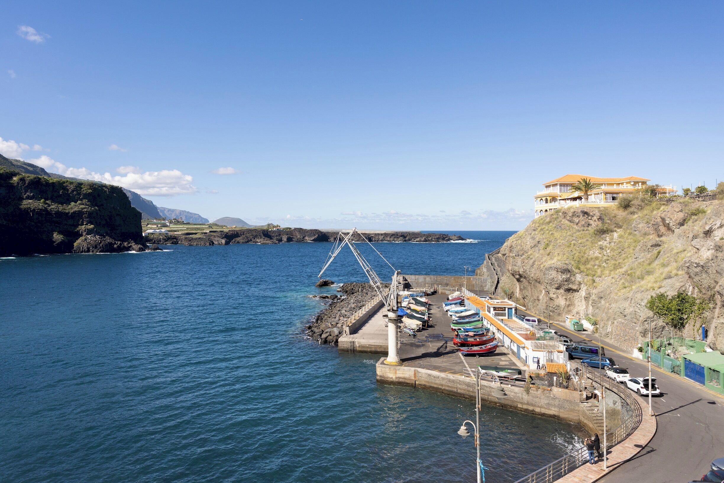 A coastal marina with boats docked, a pier, and a large building on a hillside.