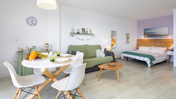 A modern, well-lit living room with a dining area, a green sofa, a bed with a wooden headboard, and a wall clock. - TenoHomes Atlantic View Icod Terrace (San Marcos)