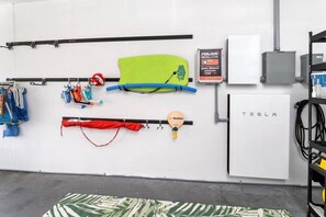 Ein Zimmer mit weißer Wand, einer Tesla-Ladestation sowie diversen Gegenständen wie einem grünen Stand-Up-Paddle, einem roten Paddel und einem blauen Kajak.