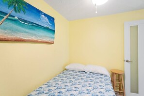 Ein Schlafzimmer mit Bett, Hocker, einer Tür und einem Wandbild mit Strandmotiv.