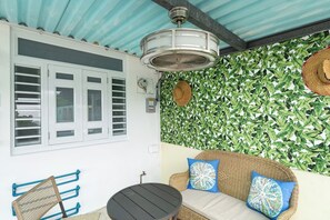Une pièce avec un ventilateur de plafond, un mur orné d’un motif végétal, un canapé en osier avec des coussins, une petite table et une fenêtre avec des volets.