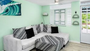 A room with a light green wall, a white sofa with patterned cushions, a ceiling fan, a window with shutters, and a painting of a wave. - Jardines Casita - 5 min walk to downtown Rincon (Ensenada)