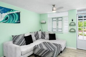 Ein Zimmer mit einer hellgrünen Wand, einem weißen Sofa mit gemusterten Kissen, einem Deckenventilator, einem Fenster mit Fensterläden und einem Gemälde einer Welle.