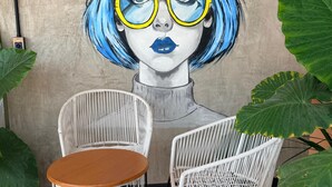 A mural with text and a stylized portrait of a person with blue hair and glasses. There are wicker chairs and a round table in front of the mural. - The coolest spot to experience the city. (Mazatlán)