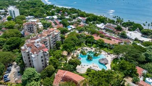 Ein Luftbild eines Resorts mit einem großen Schwimmbad, umgeben von üppiger Vegetation und einem Strand mit Booten.