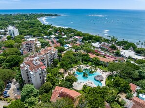 Ein Luftbild eines Resorts mit einem großen Schwimmbad, umgeben von üppiger Vegetation und einem Strand mit Booten.