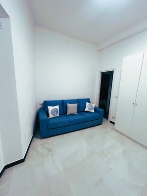 Ein Zimmer mit einem blauen Sofa, zwei Kissen, einem weißen Kleiderschrank und einem Fliesenboden.