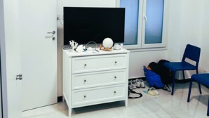 Ein Zimmer mit einem weißen Schminktisch, einem Fernseher, einem Fenster mit Jalousien, einem blauen Stuhl und einem Rucksack auf dem Boden.