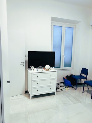 A room with a white dresser, a television, a window with blinds, a blue chair, and a backpack on the floor. - Casa Marietta - Studio Apartment, Sleeps 2 (Arenzano)