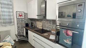 A compact kitchen with white cabinets, a stainless steel range hood, a built-in oven, and a refrigerator with a digital display.
