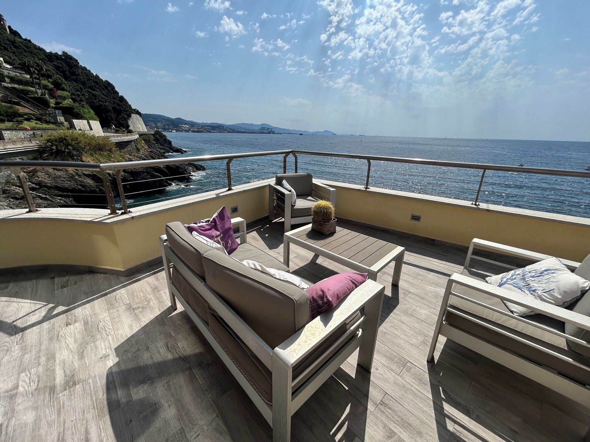 A deck with a sofa, a small table, and a chair, overlooking a body of water and a distant coastline.