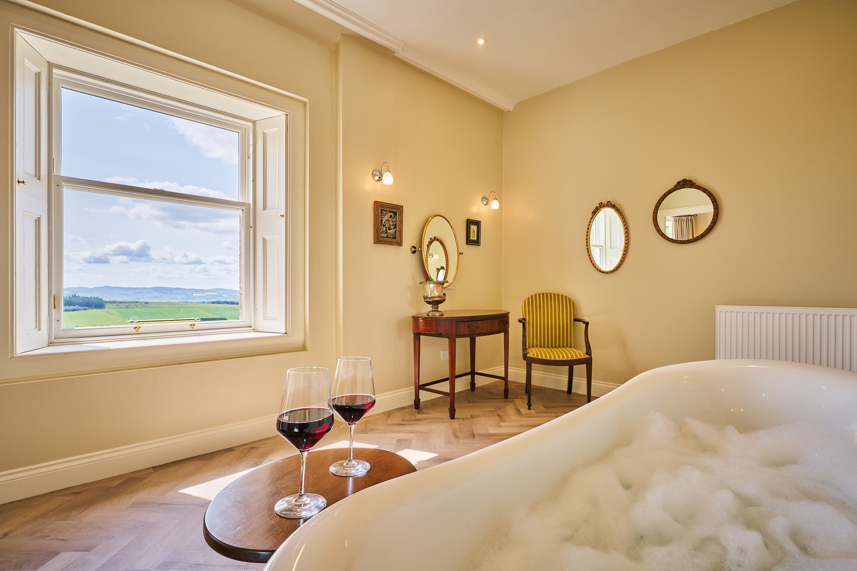 Una habitación con un ventanal grande, una bañera, dos copas de vino, una silla y una mesa.