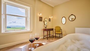 Una habitación con un ventanal grande, una bañera, dos copas de vino, una silla y una mesa.