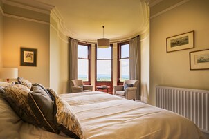 Habitación de hotel con una cama grande, dos sillas, una mesa y vistas al campo a través de ventanas amplias.