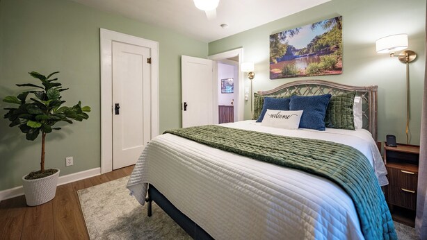 A bedroom with a bed, a ceiling fan, a wall-mounted lamp, a door, and a potted plant. - Walk to Broad Ripple & Monon Trail | Fire Pit | Family Home | Sleeps 8 (Indianapolis)