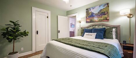 A bedroom with a bed, a ceiling fan, a wall-mounted lamp, a door, and a potted plant.