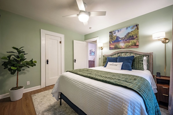 A bedroom with a bed, a ceiling fan, a wall-mounted lamp, a door, and a potted plant.