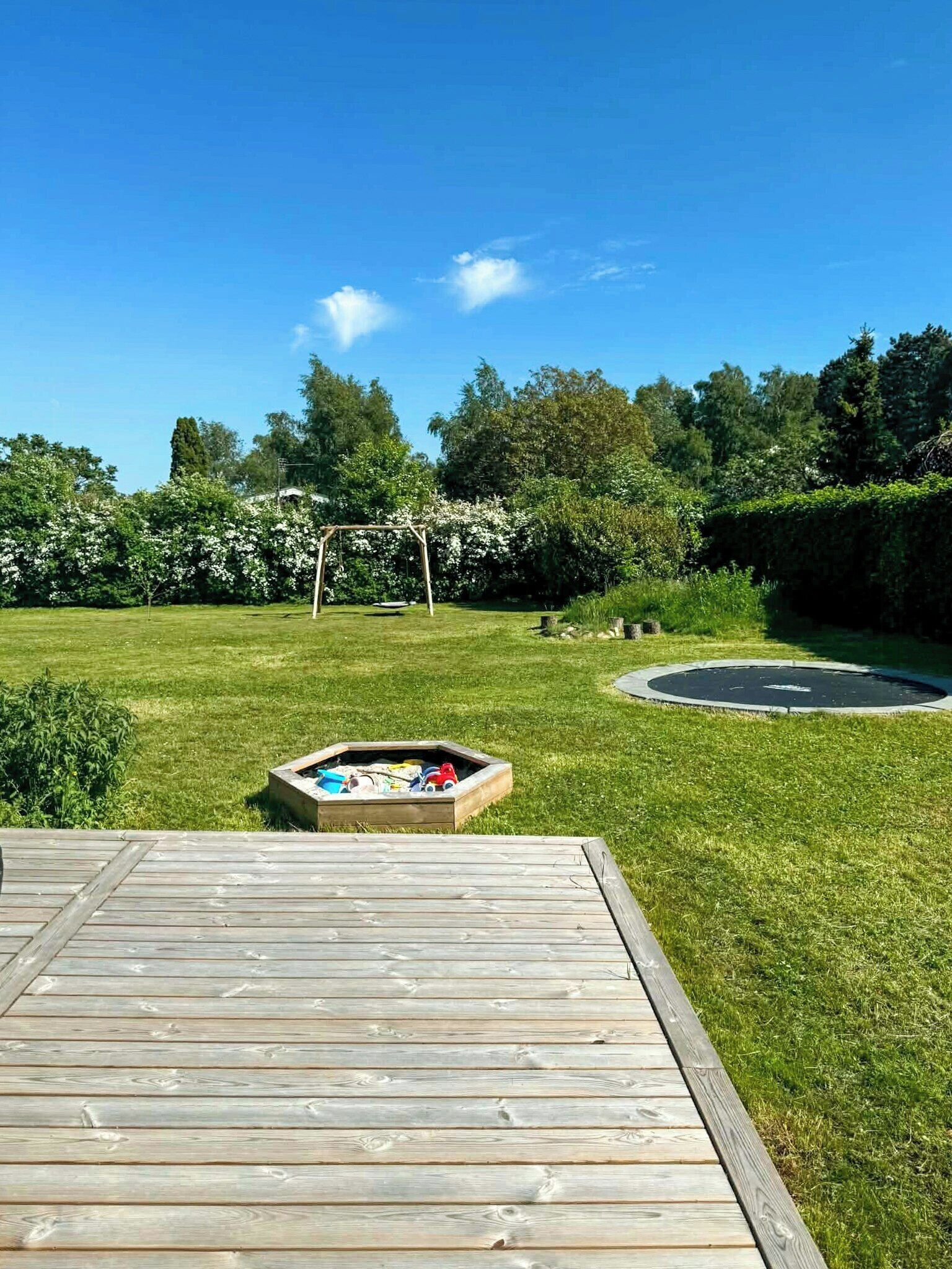 A wooden deck leading to a backyard with a sandbox, a swing set, and a circular garden bed.