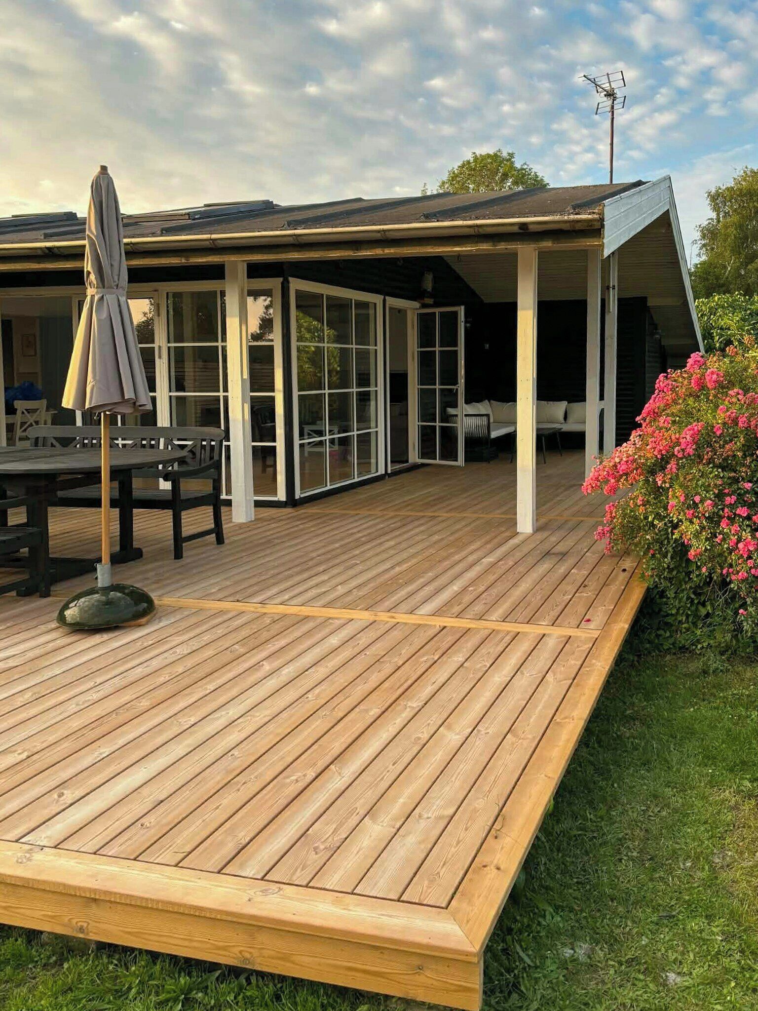 A wooden deck with a covered patio area, outdoor furniture, and a garden with pink flowers.