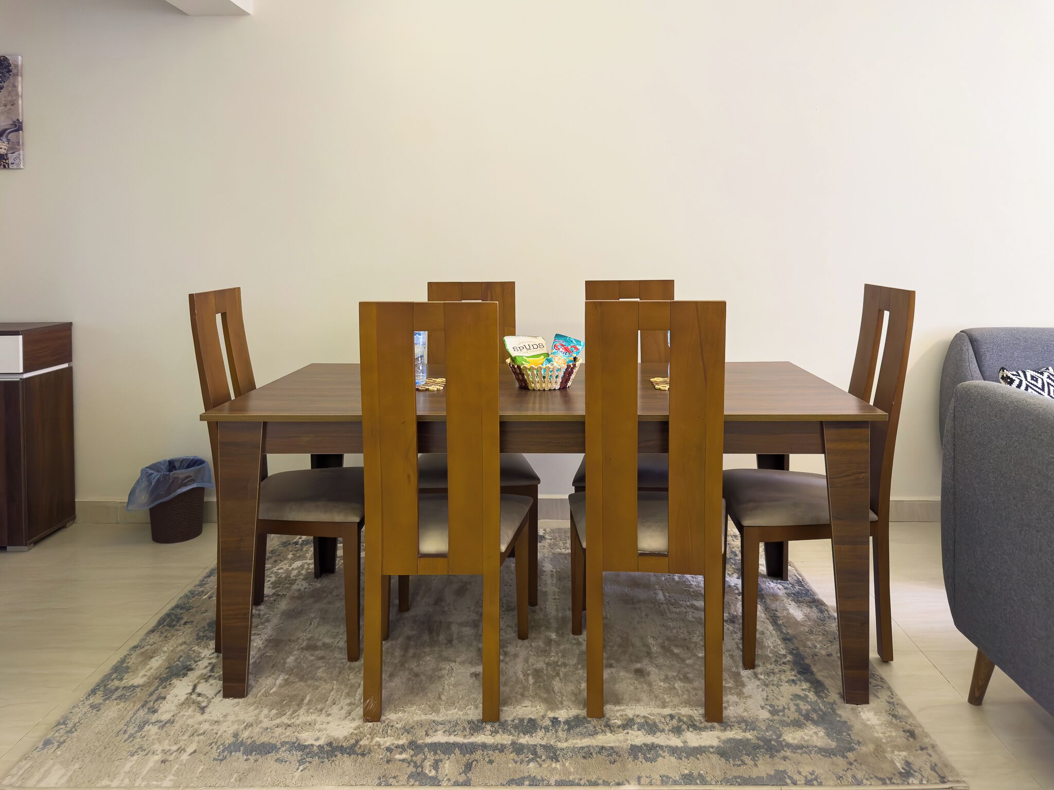 Un comedor con una mesa y sillas de madera, un cubo de basura pequeño y un sofá gris.