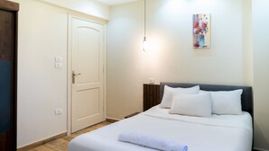 A neatly made bed with white linens and a rolled towel, a wooden floor, a white door with a handle, and a painting on the wall. - CK 803 . Spacious 2BR Apt., 2 Baths (Sheikh Zayed City)