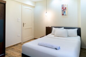 Ein ordentlich bezogenes Bett mit weißen Leinen und einem aufgerollten Handtuch, ein Holzboden, eine weiße Tür mit Griff und ein Bild an der Wand.