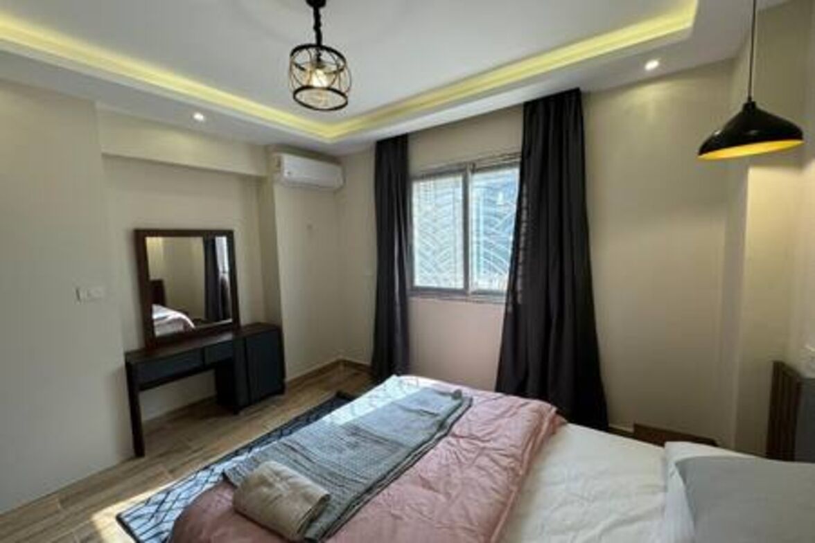 Une chambre à coucher comprenant un lit, une table de chevet, un miroir, une fenêtre avec des rideaux et un luminaire suspendu.