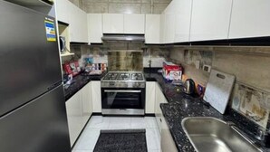 A compact kitchen with white cabinets, a stainless steel refrigerator, a built-in oven, and a black countertop. - CK 903 . Spacious 2BR Apt., 2 Baths (Sheikh Zayed City)