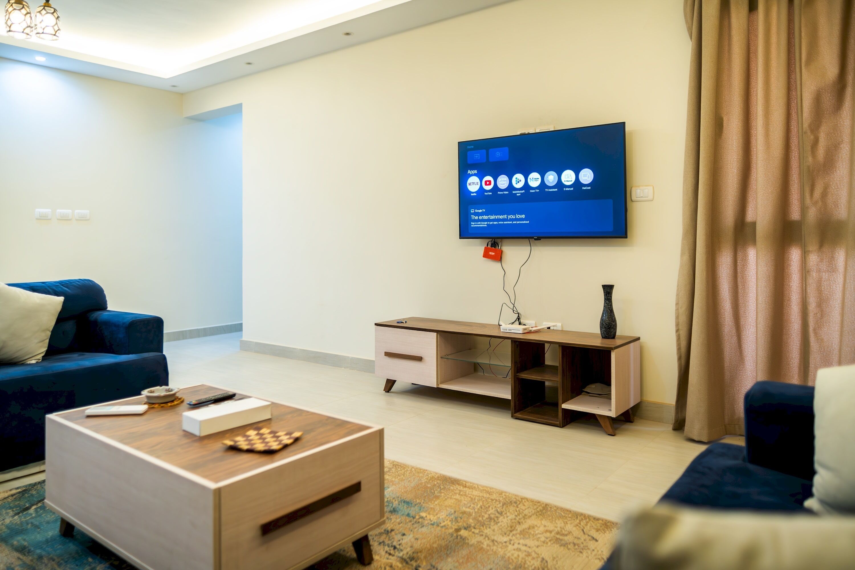 A modern living room with a flat-screen TV mounted on the wall, a wooden coffee table, a blue sofa, and a small TV stand with a vase.