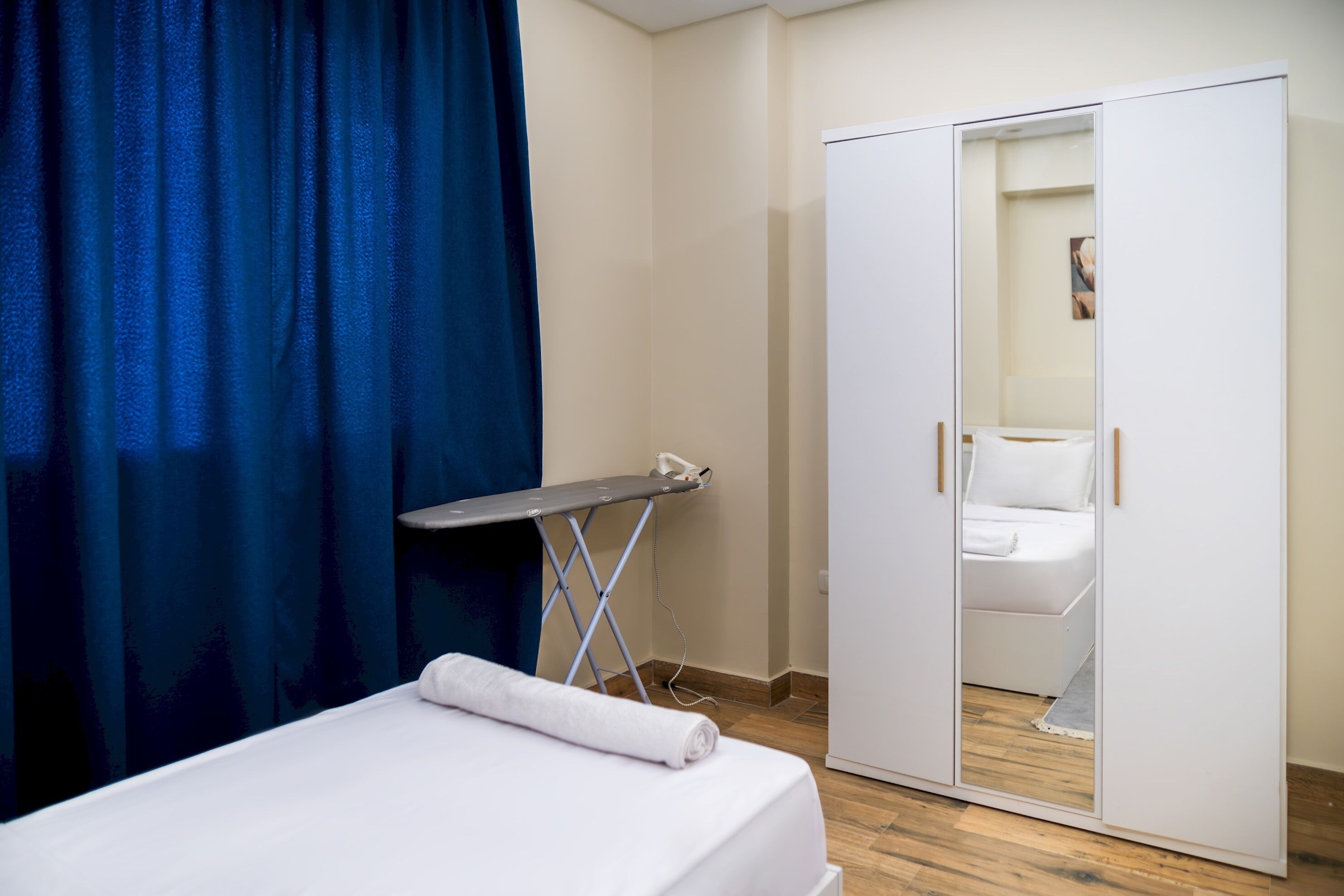 Una habitación de hotel con cama, tabla de planchar, armario y cortina azul.