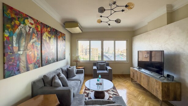 A living room with a grey sofa, a round coffee table, a TV on a wooden cabinet, and a large window with a city view. - 1406.  Elegant 2BR Apt | Sun Terrace | Ensuite (Almazah)