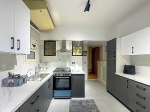 A modern kitchen with a gas stove, oven, and a microwave. There is a sink with a faucet, and cabinets with handles. A window with blinds is visible on the left wall. - 1406.  Elegant 2BR Apt | Sun Terrace | Ensuite (Almazah)