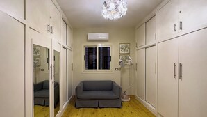 A room with a grey sofa, wooden flooring, white cabinets, a ceiling light fixture, and a window with blinds. - 1406.  Elegant 2BR Apt | Sun Terrace | Ensuite (Almazah)