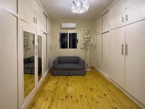 A room with a grey sofa, wooden flooring, white cabinets, a ceiling light fixture, and a window with blinds. - 1406.  Elegant 2BR Apt | Sun Terrace | Ensuite (Almazah)