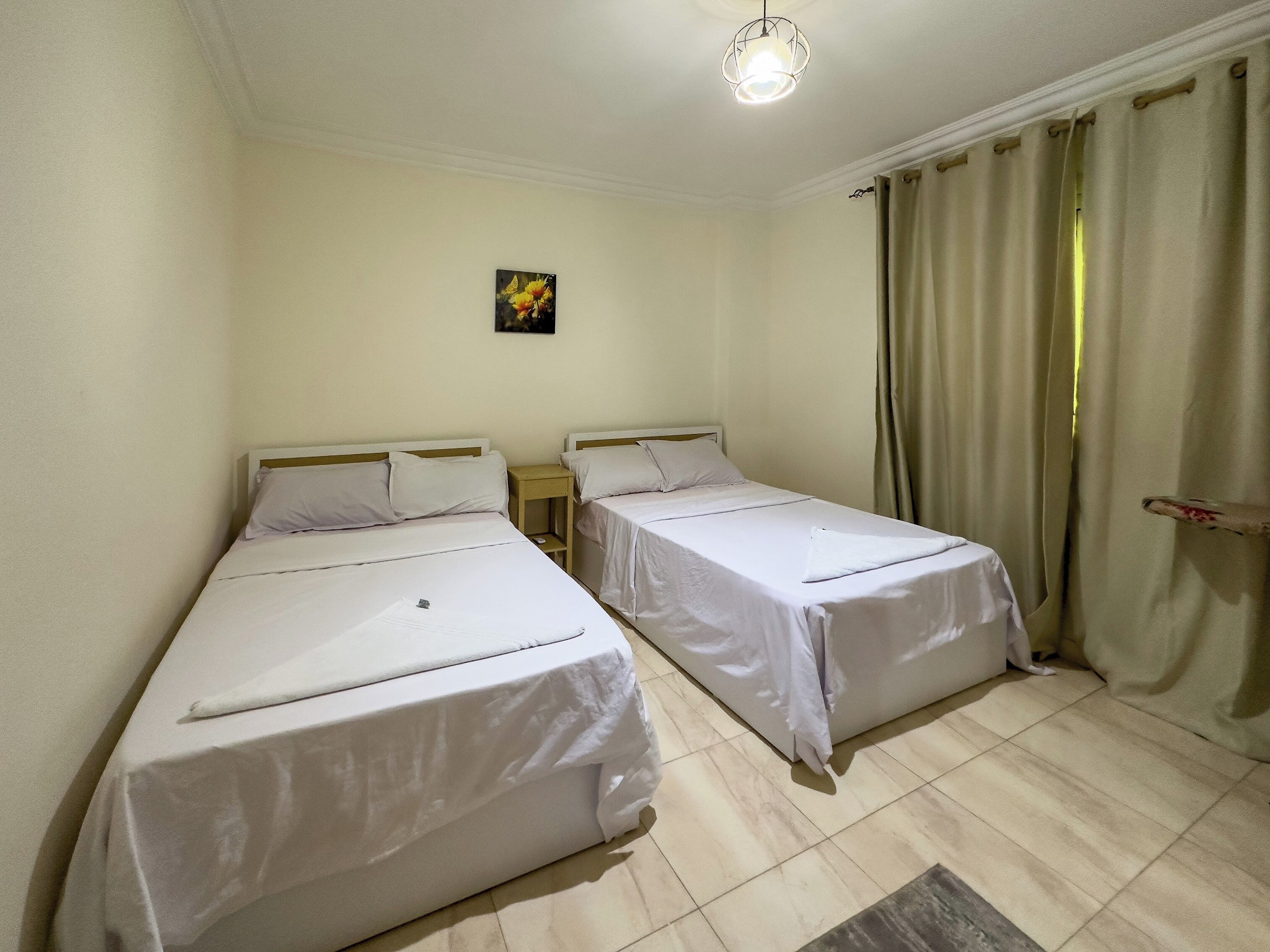 A room with two single beds, a small table, a wall-mounted light fixture, and a window with curtains.