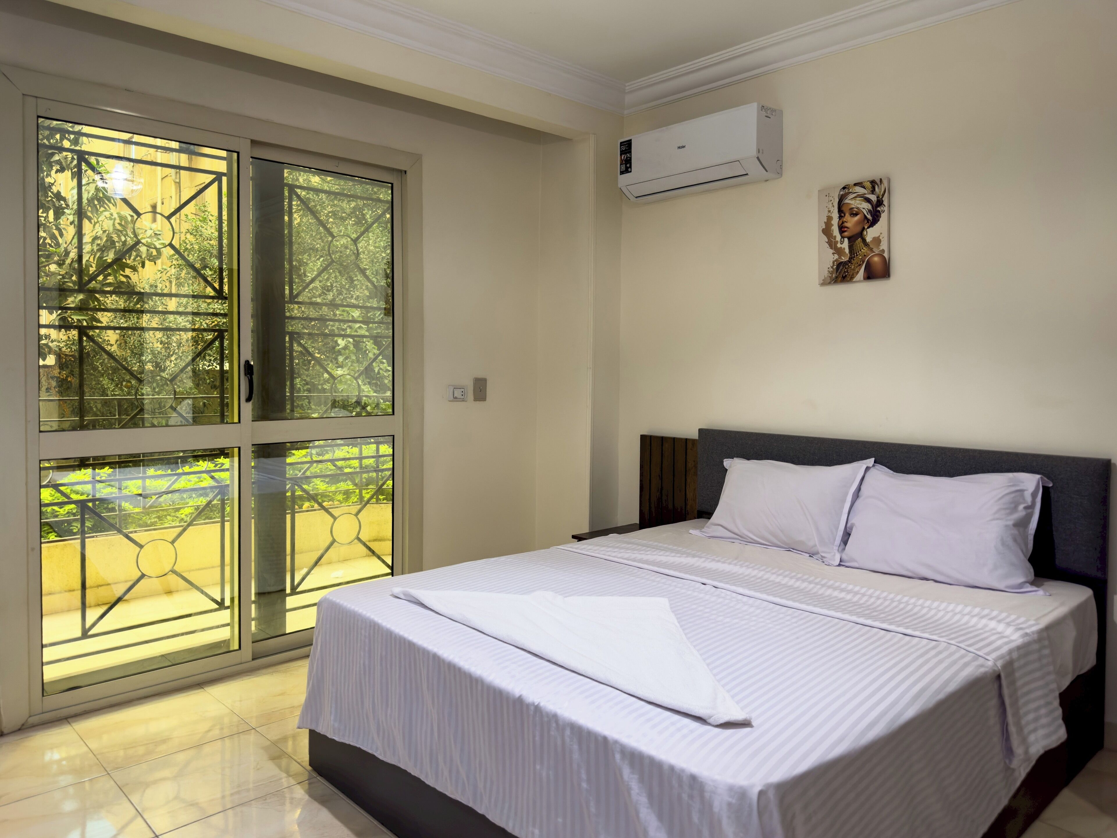 A hotel room with a bed, a window with a view of greenery, and an air conditioning unit on the wall.